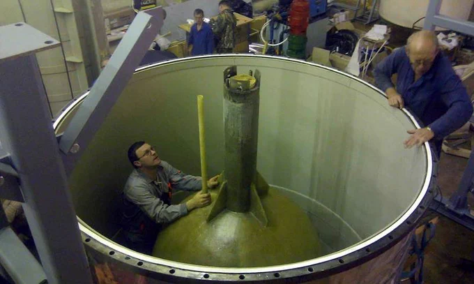 A large tank with a metal sphere with a rod coming out of the top of it inside of the tank, which is part of the BEST experiment apparatus. A researcher (a white man in a gray shirt) is standing next to the sphere and another researcher (a white man in a blue shirt) is standing outside of the tank.