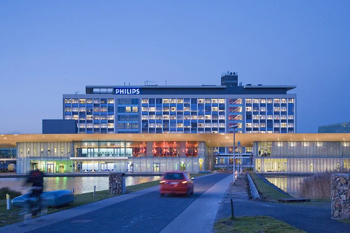 A wide shot of the High Tech Campus Eindhoven with moody lighting in the near-evening with water in front of the buildings while a car is driving by.