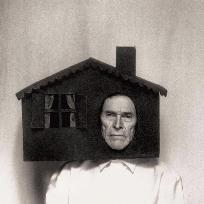 A man in a photo shoot with a small wooden house on his head, face sticking through the entrance oval