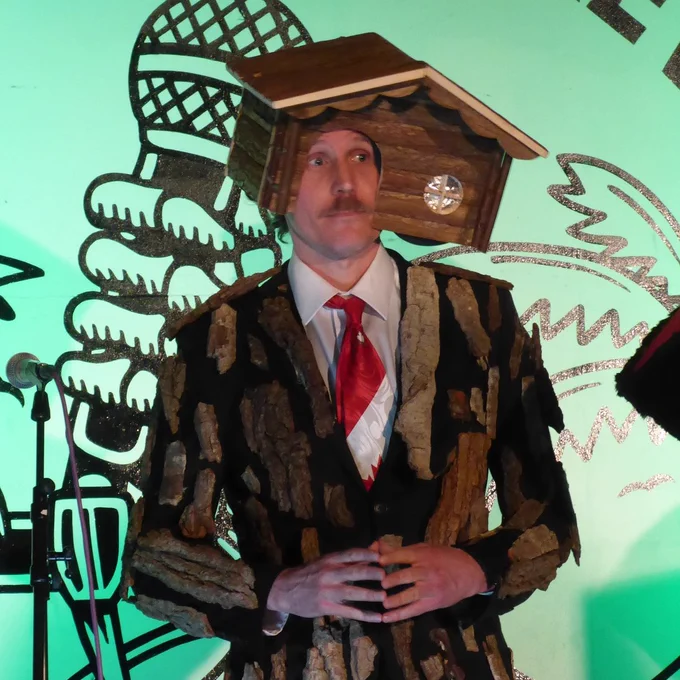 A man onstage with a small wooden house on his head, face sticking out of the door arch