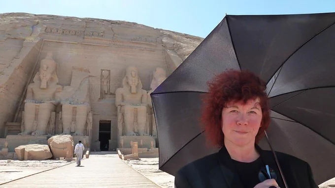 Professor Joann Fletcher stands with a black umbrella outside the temple of Abu Simbel, Egypt. The temple’s frontage of colossal statues dominate the background.