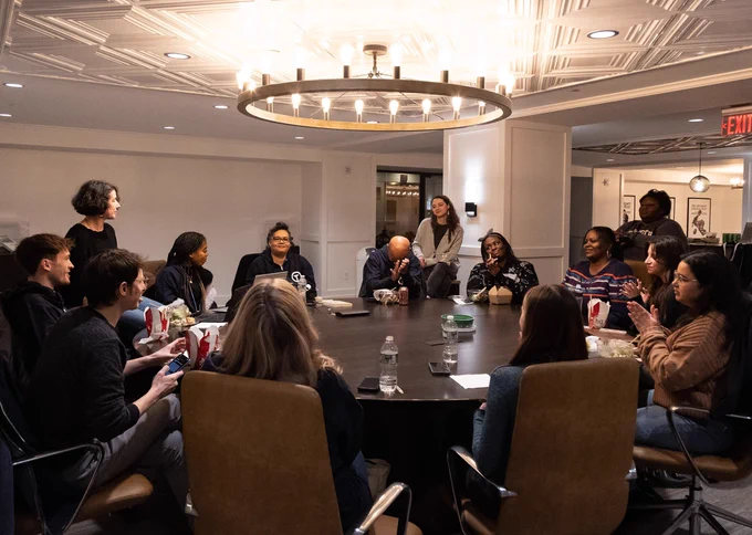 A group of individuals sitting and standing around a large round table in a conference room, engaging in discussion. The room has a modern design with a prominent overhead light fixture.