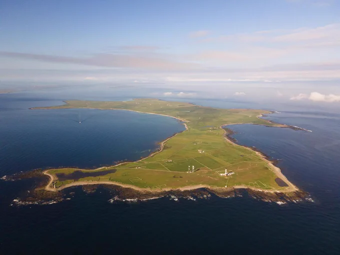 Aerial photograph of North Ronaldsay in the sunshine, with a deep blue sea, pale blue sky. The lighthouse and beacon on the north end are visible. Taken by @papayranger.