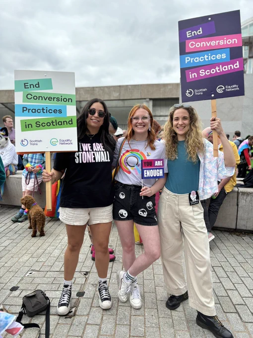 Denisha, Rebecca and Eleanor standing in front of Scottish parliament holding signs that read “end conversion practices in Scotland.”