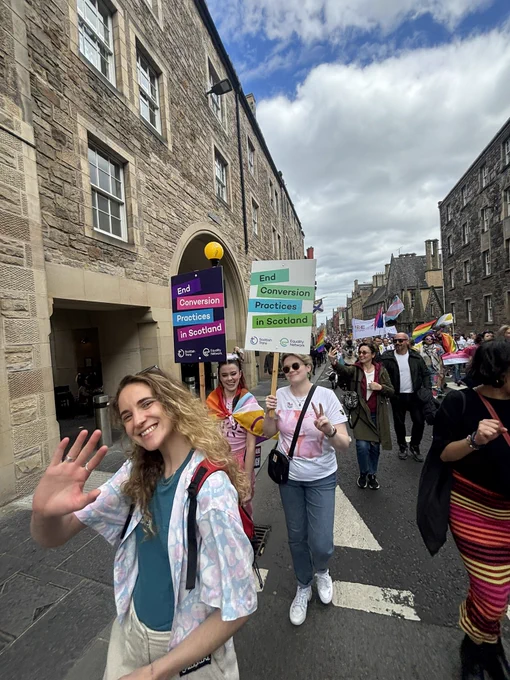 Eleanor, Erin and Orla marching. Eleanor is waving at the camera and Erin and Orla are holding signs that read “end conversion practices in Scotland”