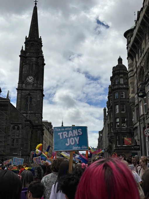 Photo of the backs of people’s heads on the pride march. Prominent sign reads “this is what trans joy looks like”
