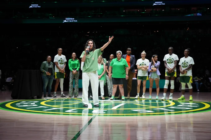Sue Bird with a microphone standing in front of the Storm owners and members of the Storm 25