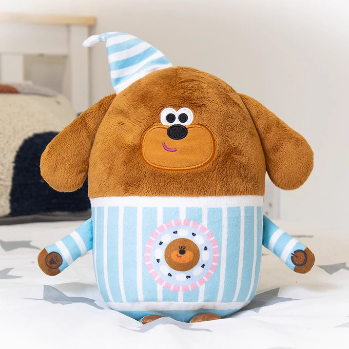Hey Duggee sleepytime plush toy (Duggee wearing blue striped pyjamas and a night hat) sits on a bed. 