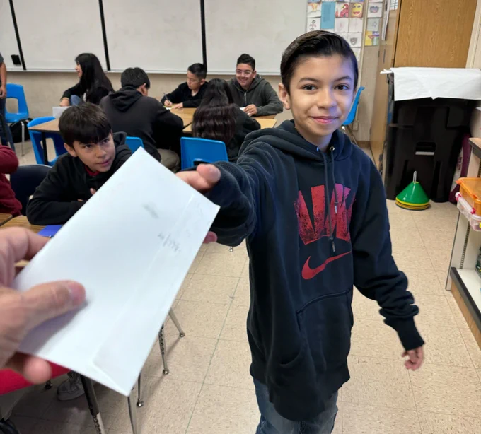 A student in a navy blue hoodie with 'Nike' written in red on it is smiling as his teacher hands him a letter from his pen pal.