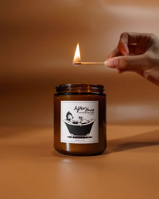 A struck match atop Answer in Progress's candle called "After Hours". The candle label features a woman relaxing in a bath with a drink in hand. 
