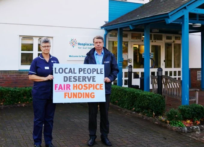 Hospiscare nurse Julie and CEO Andrew hold a sign reading "local people deserve fair hospice funding" in front of the hospice building.