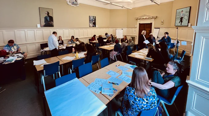 A wide angled image of a room of people doing the workshop. They have loads of paper on the tables.