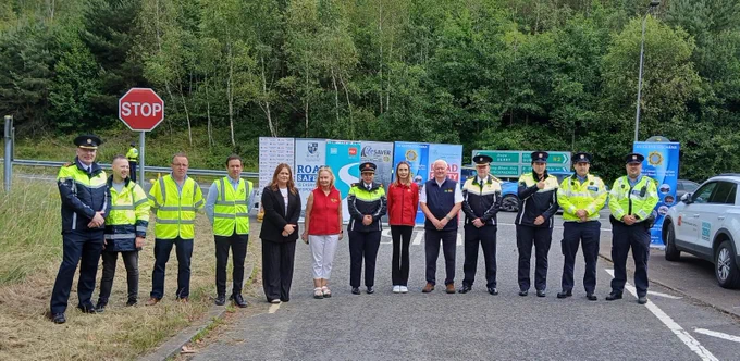 A group of 13 people standing along a road with a stop sign behind them, a number are Gardai. There are banners behind the group and tall trees along the road in the background.