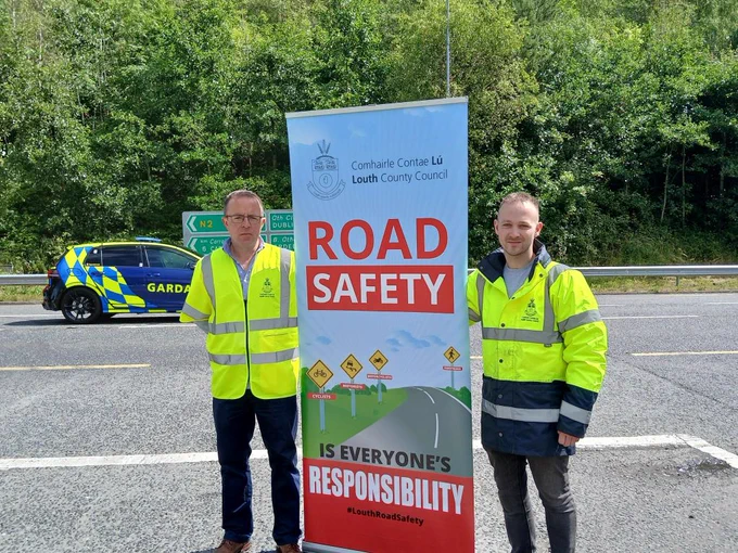 Enda Murphy, Senior Executive Engineer, Louth County Council and Aaron Lynch, Assistant Engineer Louth County Council hold a 'Road Safety' banner on a main road with a Garda patrol car in the background and trees along the road.