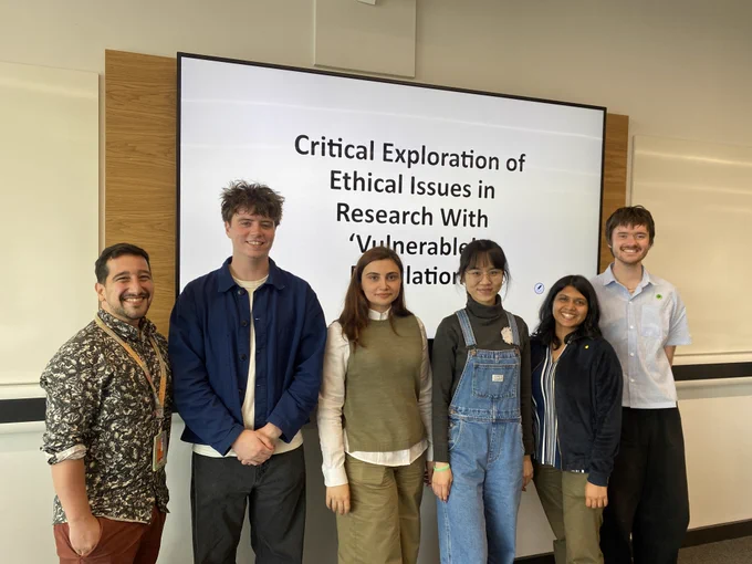 (from left to right): Antonios, Tom, Turana, Hsiao-fang, Ankita and Charlie stand in front of a screen with the title of the symposium: Critical Explorations of Ethical Research With 'Vulnerable' Populations.