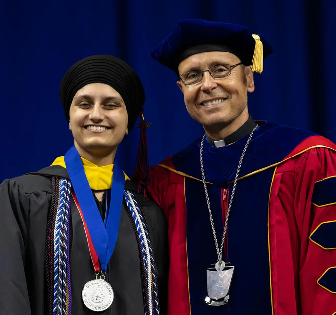 A young woman in a headscarf wearing a graduation robe and cords and a medal around her neck smiles next to an older man wearing a priest's collar and ornate university regalia.