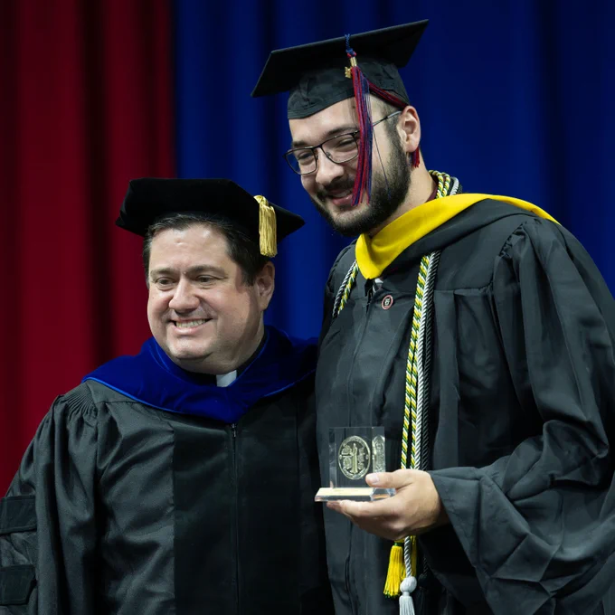 A young man with glasses wearing a graduation robe, cap, and cords holds up a glass award and smiles next to a man wearing a priest's collar and university regalia.