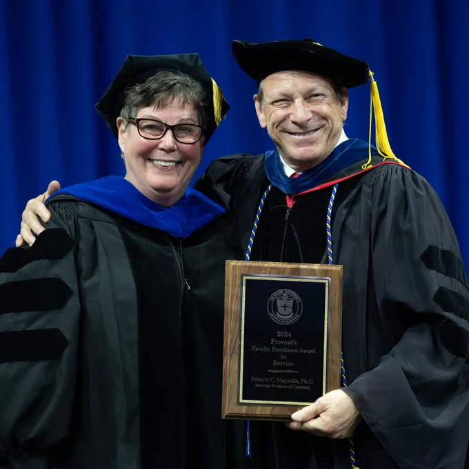 A man and a woman with glasses both wearing university regalia smile into the camera and the man proudly holds up a plaque.