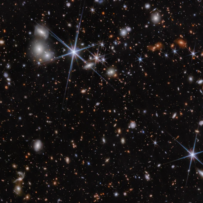 Hundreds of stars and galaxies in varying shapes, sizes and colors on a black background. Two of the largest, most prominent stars can be found in the top left quadrant and near the bottom right edge, respectively. They are tinged with blue and have an eight-point diffraction spike pattern. Near the center of this image is the ZS7 system, made up of a merger between two galaxies and their black holes. The ZS7 system is not easily seen by the naked eye in this image, as it looks like very small red dots.