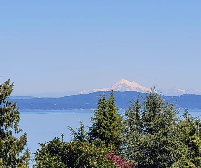Photo of the snow-capped volcano, Koma Kulshan (Mt. Baker) as seen from southern Vancouver Island. Green trees, distant islands and a volcano, 125 km away.