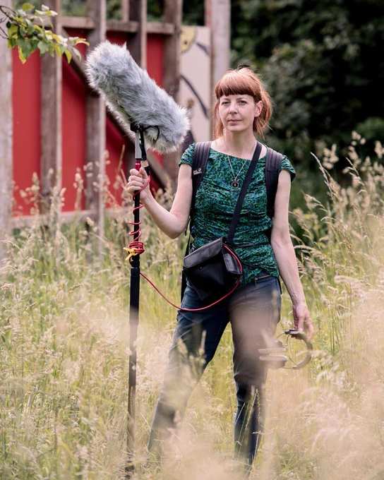 Artist Fiona Hamilton stands in a grassy area holding a microphone boom and wearing a sound recording kit