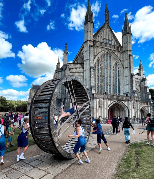 A group of performers push a giant wooden whenever in front of a crowd towards Winchester cathedral. A woman is performing inside the wheel.