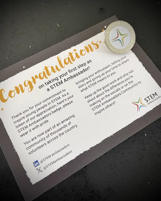 A congratulatory card from STEM Ambassadors on a black surface, featuring elegant gold and red text and a gold star-shaped badge with "Ambassador" on it. The card reads: "Congratulations on taking your first step as a STEM Ambassador" and includes social media handles @STEMAmbassadors.