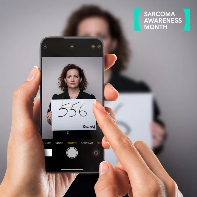 Photo from the perspective of someone taking a photo with a phone. The phone is in the foreground and the subject is a white woman called Philippa holding. a card in front of her with '556' written. 