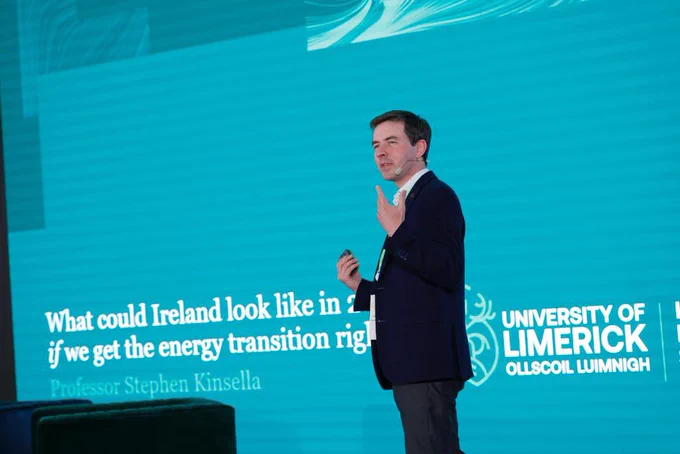 A photograph of a man in  dark blue suit jacket, white shirt and blue trousers. He is giving a speech and is stood in front of a blue screen with white text on it reading: "What could Ireland look like if we get the energy transition right? Professor Stephen Kinsella". The University of Limerick logo is also visible in the bottom right of the screen
