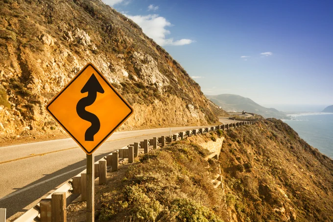 Curvy road sign along a coastal highway with ocean and hills in the background on a sunny day.