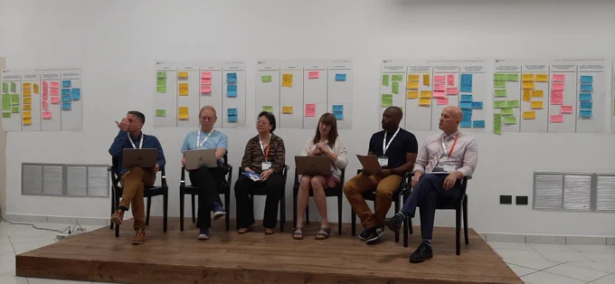 Panel of six people (mixed gender, ethnicity and nationality) seated on a podium with a wall featuring multiple post-it notes behind 
