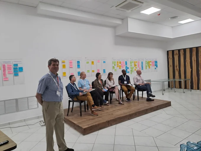 Panel of six people (mixed gender, ethnicity and nationality) seated on a podium with a wall featuring multiple post-it notes behind and a man in the foreground