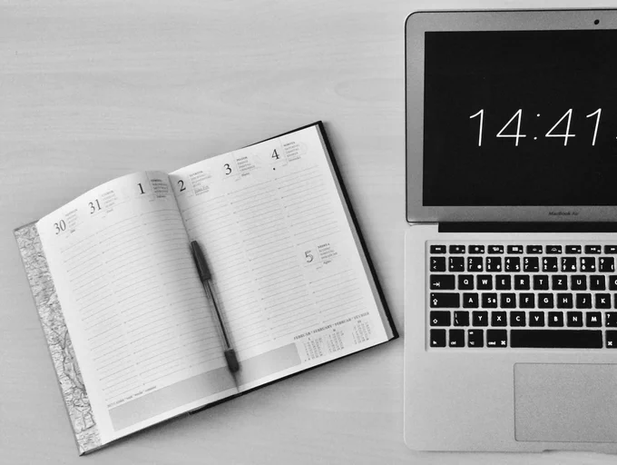 Photo of a planner book with a pen and a laptop stating the time. 