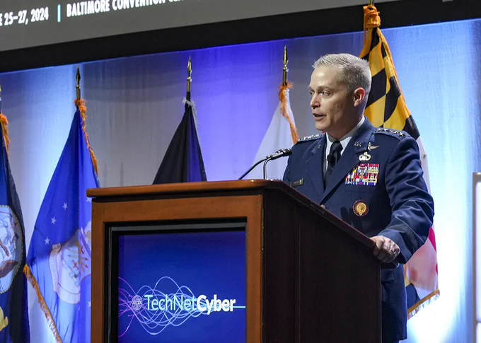 A man in uniform stands speaking behind a podium with flags in the background.