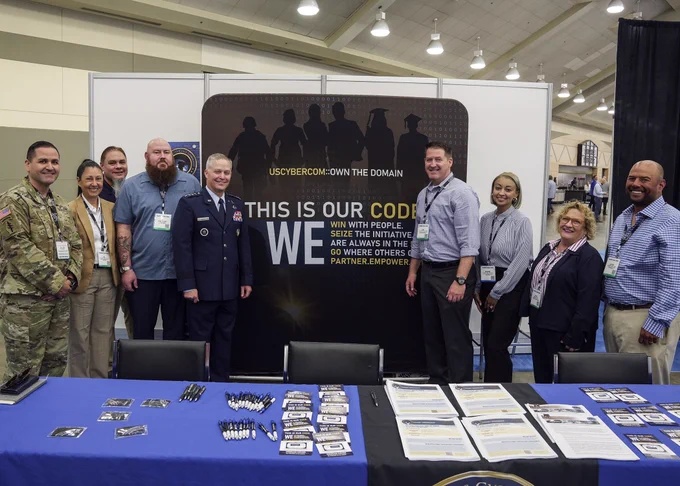A group of men and women pose around a sign that says "USCYBERCOM -Own the Domain." In front are two tables covered in pamphlets, pens, and other swag.