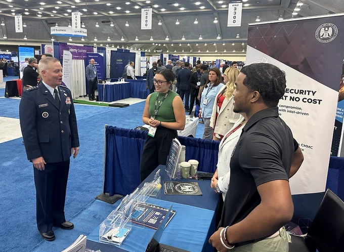 A man in uniform stands in front of a booth talking to a group of people on an exhibition floor.