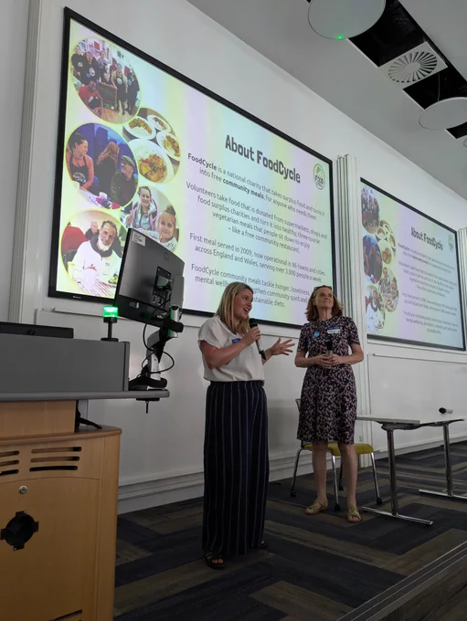 Two women smiling and speaking in front of a presentation slide titled About FoodCycle