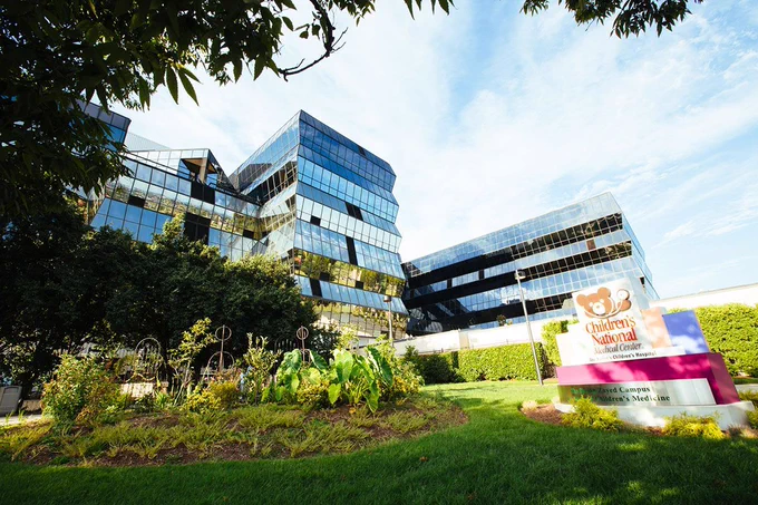 A aospital building with slanted window configuration behind a green park and sign with a brown teddy bear.