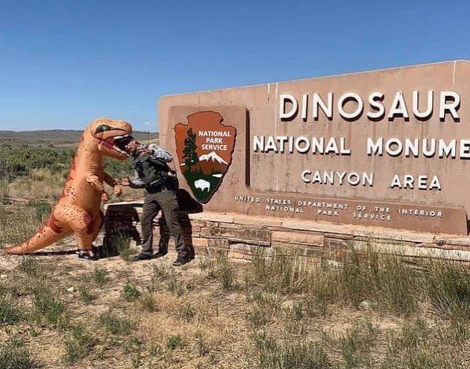Large inflatable dinosaur and a ranger have an awkward moment in front of park sign.