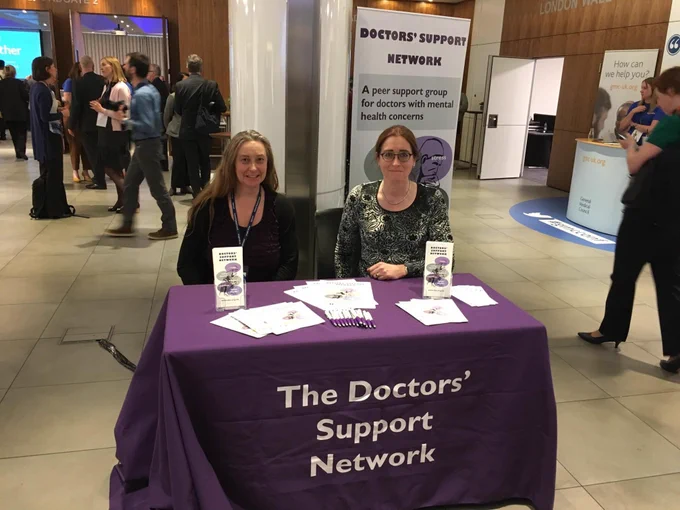 Two women sitting behind table with purple cloth and text ‘The Doctors’ Support Network’
