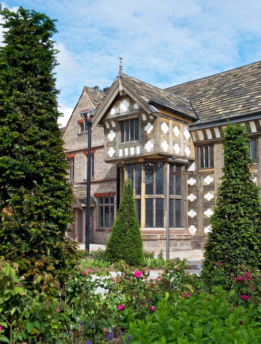 The outside of Ordsall Hall's oriel window seen through yew trees and rose bushes.