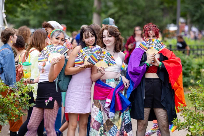 Four people are standing and holding Pride fans that read, "Make Pride Accessible!" They are wearing Pride clothing and have a Pride flag.