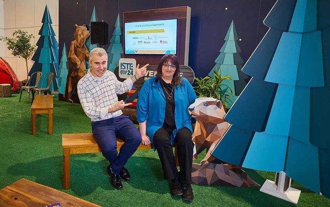 Two individuals seated on a bench in an indoor setting with a forest-themed display including stylized trees and an animal sculpture. The backdrop features a screen with text thanking sponsors, including names like Cisco, AWS, Intel, among others.
