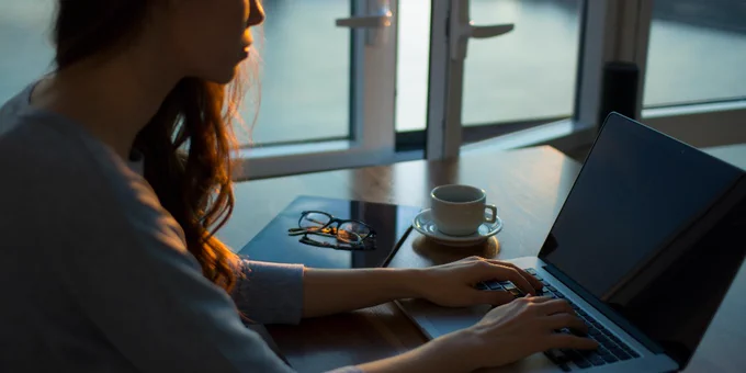 Stock image of woman typing on a laptop in low lighting. A pair of glasses, a coffee mug and an open window are visible in the background.