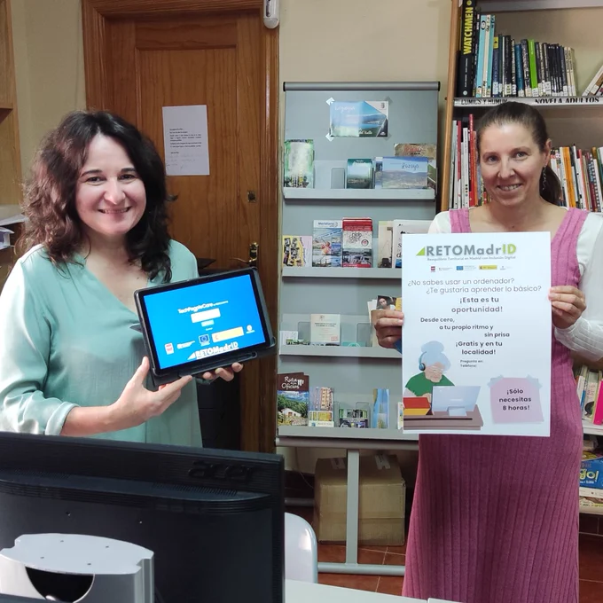 Fotografía del interior de la Biblioteca de Lozoya con dos mujeres sonriendo a cámara. Una sosteniendo una tablet en la mano, con la portada del curso de RETOMadrid en ella, y la otra sosteniendo un cartel del proyecto animando a la población a apuntarse al curso. Fondo de librerías y folletos de la biblioteca.