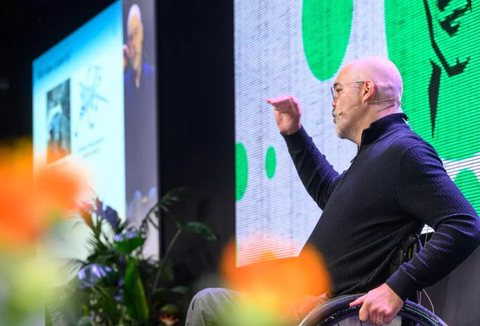 A man in a wheelchair (Jochen Guck) is holding a speech at a stage and gesturing with his hand. 