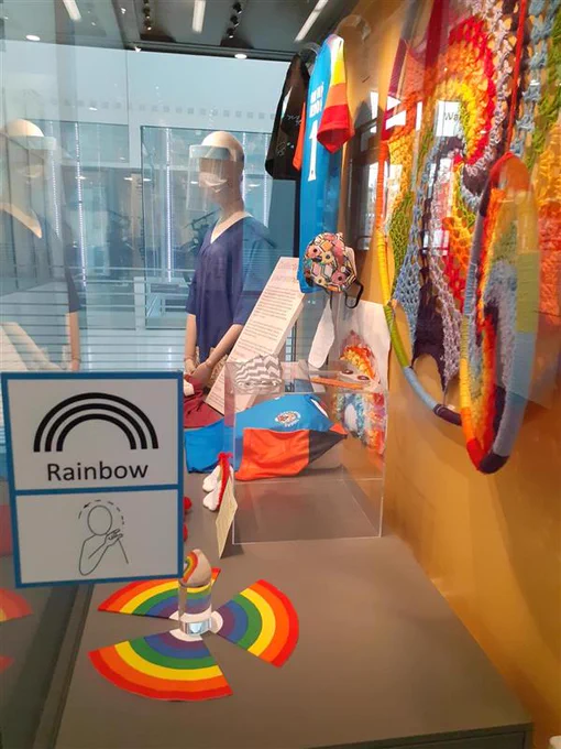 A display case containing items relating to the local COVID-19 response, including medical scrubs, face masks and rainbows. There is also the Makaton sign for ‘rainbow’ displayed on the side.