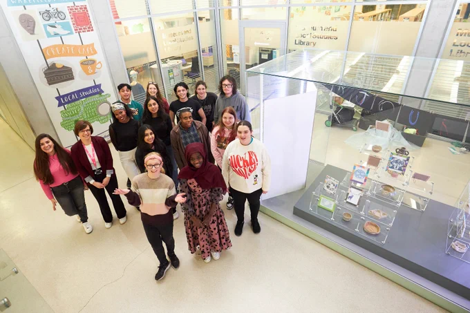 The 12 members of the Wakefield: Where We Are youth group with group facilitators grinning at the camera, next to their newly installed ‘Our Journey’ display case at Wakefield One