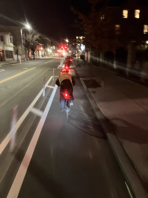 A photo at night of a bike lane with flexposts on the side. In the bike travel lane there are approximately 4 or 5 cyclist visible; there are no cars visible on the road next to the cyclists.