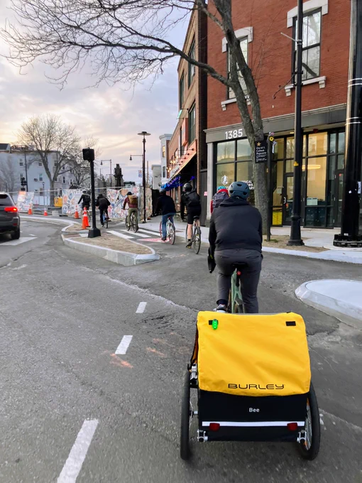A line of 8 bikers enter into an off-street cycletrack next to a construction area. One is pulling a bright yellow Burley trailer.